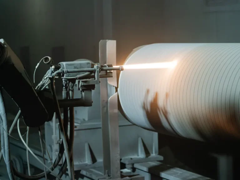 A robotic arm precisely spraying tungsten onto a rotating corrugated roll in a manufacturing setting.